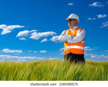 Confident Man Engineer. Engineer In White Helmet On Field. Employee Engineering Bureau Against Background Sky And Grass. Man Works In Engineering Business. Concept Of Site Selection For Construction