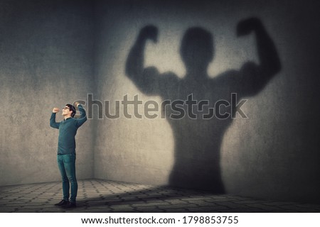 Confident man flexing muscles imagine super power as casting a shadow of muscular bodybuilder showing biceps. Strong person facing his fears. Personal development, inner strength, motivation concept.