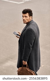 Confident man glances back while holding his phone and a brown bag, standing on the street.