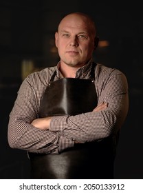 Confident Man In The Leather Apron Folded His Arms Standing On Dark Background Looking At The Camera. Portrait Of Handicraft Business Owner