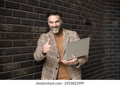 A confident man in a plaid coat shows a thumbs-up gesture while holding a laptop, leaning against a dark brick wall in an urban setting. He looks pleased. - Powered by Shutterstock