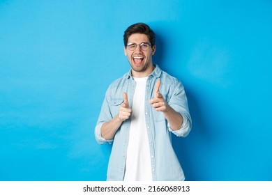 Confident man saying congrats, winking and pointing at you, standing pleased over blue background and smiling - Shutterstock ID 2166068913