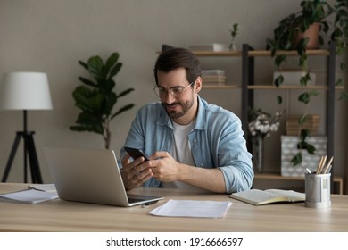 Confident man sitting at desk taking break in work with electronic documents on laptop to make answer telephone call. Smiling young guy freelancer synchronize data between home computer and smartphone - Powered by Shutterstock