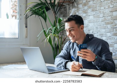 Confident man sitting at desk taking break in work with electronic documents on laptop to make answer telephone call. Smiling young guy freelancer synchronize data between home computer and smartphone