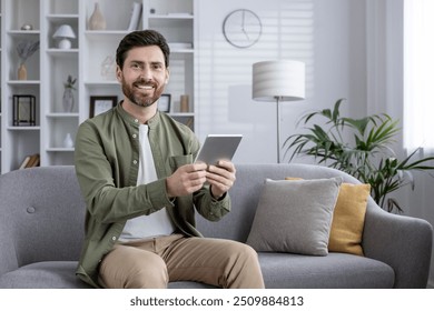 Confident man wearing green shirt sits comfortably on sofa using tablet. Living room setting conveys modern lifestyle and relaxation. No background description included in keywords. - Powered by Shutterstock