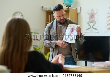 Confident mature biology teacher showing paper with frog picture to student while making presentation and explaining its internal structure