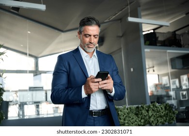 Confident mature business man ceo wearing blue suit standing in office using cell phone. Mid aged businessman professional executive holding mobile smartphone working, texting messages on tech device. - Powered by Shutterstock