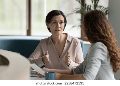 Confident mature business professional woman talking to younger female colleague at office table, speaking, gesturing, teaching, explaining work tasks. Elder mentor training intern