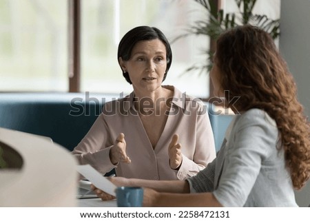 Confident mature business professional woman talking to younger female colleague at office table, speaking, gesturing, teaching, explaining work tasks. Elder mentor training intern Foto stock © 