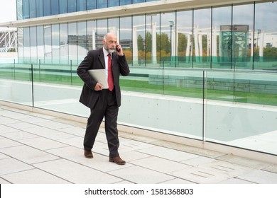 Confident Mature Businessman Talking On Cellphone On His Way To Office. Elderly Man In Formal Suit And Tie Walking Outside In City. Business Communication Concept