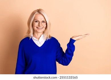 Confident mature businesswoman with glasses presenting, blue jumper against beige background - Powered by Shutterstock