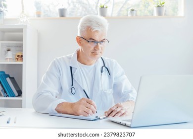 Confident mature caucasian senior male doctor making notes while using laptop for telemedicine with patients at hospital. GP make online video call consult patient. Online remote medical appointment