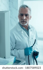 Confident Mature Doctor In A White Coat Looking At The Screen Of A Monitor And Holding An Endoscope