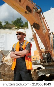 Confident Mature Foreman In Eyeglasses, Reflection Vest, Uniform And Hardhat Keeping His Arms Crossed On Chest Against Excavator