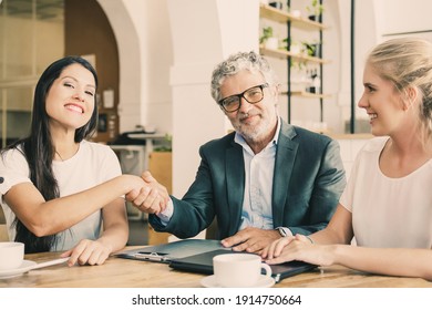 Confident Mature Investor Shaking Hands With Young Entrepreneurs. Man And Women Meeting Over Cup Of Coffee At Co-working. Medium Shot. Partnership And Investment Concept