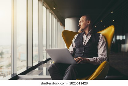 A confident relaxed successful mature man entrepreneur sitting with a laptop on an orange armchair and thoughtfully looking aside on an urban skyline outside the window; a copy space place on the left