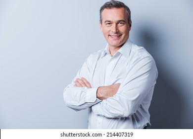 Confident Mature Man. Portrait Of Confident Mature Man In Shirt Looking At Camera And Smiling While Keeping Arms Crossed And Standing Against Grey Background