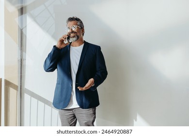 Confident mature man in stylish attire engaging in a pleasant phone conversation indoors with natural light casting shadows around him, conveying a sense of business, communication, and ease. - Powered by Shutterstock