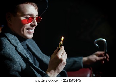 Confident Mature Man In Stylish Expensive Clothes And Glasses Sits In A Vintage Armchair And Plays With A Lighter On A Black Background. Gangster Man, Mafia. 