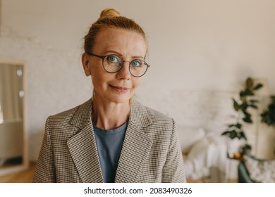Confident mature middle-aged woman family counselor in round-shaped glasses and plaid business jacket posing against home interior, looking at camera with happy, positive face expression