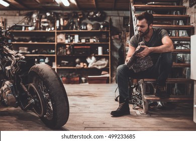Confident mechanic. Confident young man holding rag and looking at motorcycle while sitting near it in repair shop 