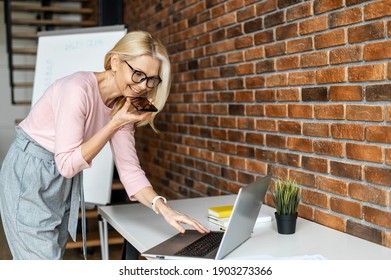 Confident Middle Aged Businesswoman In Glasses Using Voice Recognition Function On A Smartphone. Modern Female Entrepreneur Looking At The Laptop, Sending A Vocal Message Via Online Messenger