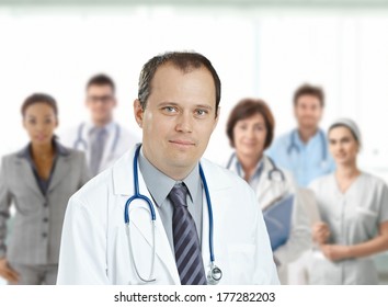 Confident Middle Aged Male Doctor Looking At Camera, Smiling, Medical Team In Background.