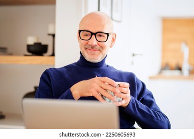 Confident middle aged man using laptop and having video call while working from home. Confident male drinking tea while sitting at table at the kitchen. Home office.