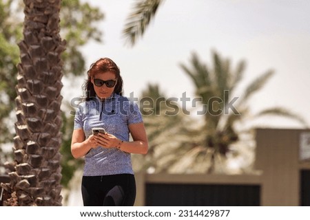 Confident middle aged woman in sportswear and sunglasses using mobile phone, looking at cell phone. Palm trees at backdrop. Healthy sports lifestyle and leisure activity concept. Copy ad text space