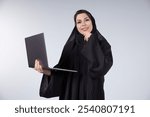 Confident Middle Eastern woman wearing a hijab and abaya, smiling while holding a laptop and thoughtfully resting her hand on her chin. Professional, modern businesswoman in a white background setting