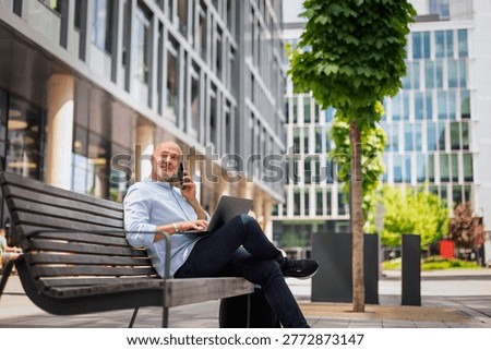 A confident middle-aged man dressed in smart casual sitting on a bench in business district. Professional businessman using smartphone and laptop for work. 