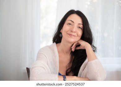 A confident middle-aged woman with long black hair, smiling in a cozy white sweater, in a well-lit room, expressing calmness concept - Powered by Shutterstock