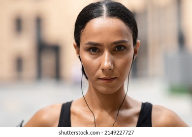 Confident Millennial Arab Female Athlete Brunette In Headphones Look At Camera On Building Background, Close Up. Workout Outdoor, Modern Training, Sports In City, Body Care During Covid-19 Quarantine