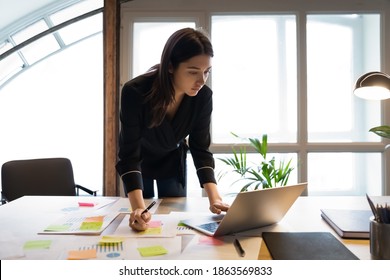 Confident millennial indian female business analyst financial advisor preparing statistic report studying documents on work desk, browsing information online using pc, writing out notes on paper sheet