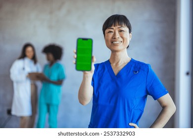 A confident nurse in blue scrubs displays a smartphone with a blank green screen. Medical professionals in the background create a healthcare setting, highlighting teamwork and technology. - Powered by Shutterstock