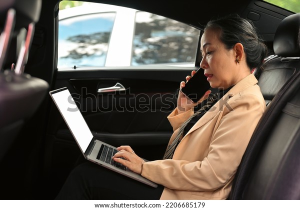 Confident old Asian executive woman in\
formal clothes concentrating on screen of laptop and interacting\
with smartphone while sitting in luxurious\
car.