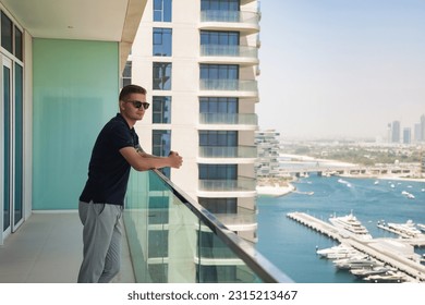 Confident pensive young man in sunglasses on skyscraper balcony with view of Dubai UAE, looking away. Attractive guy pondering on terrace of tower block. Recreation and leisure activity. Copy ad space - Powered by Shutterstock