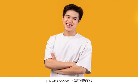 Confident Person. Portrait Of Smiling Asian Guy With Folded Arms Looking At Camera, Wearing White Shirt, Posing Isolated Over Orange Studio Background. Happy Casual Male Teenage Model Laughing