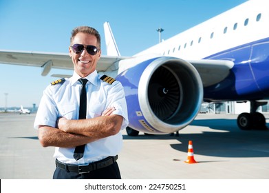 Confident pilot. Confident male pilot in uniform keeping arms crossed and smiling with airplane in the background 