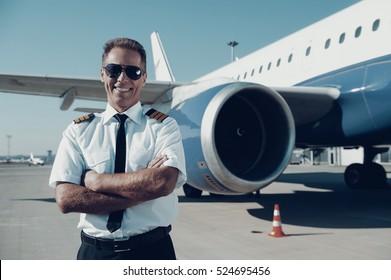 Confident pilot. Confident male pilot in uniform keeping arms crossed and smiling with airplane in the background