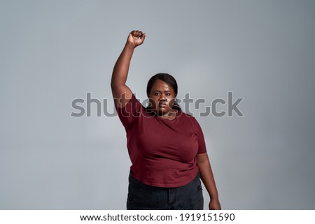 Confident plus size young african american woman in casual clothes looking at camera, standing with fist raised isolated over gray background. Social issues, protest concept. Front view