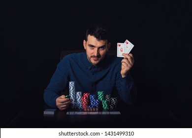 Confident Poker Player Seated At The Casino Table Showing His Winning Cards Over Black Background. Gambling Tournament Winner Success Concept. Chips And Dices Over Dark Surface.