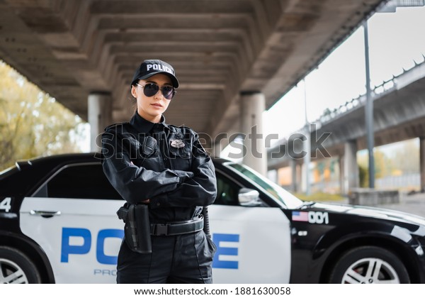 confident policewoman
with crossed arms looking at camera near patrol car on blurred
background on urban
street