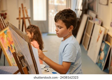 Confident portrait of a Caucasian handsome teenage school boy in blue casual shirt, holding a paintbrush and drawing picture on canvas, while learning fine art in creative art workshop - Powered by Shutterstock