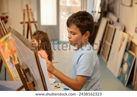Confident portrait of a Caucasian handsome teenage school boy in blue casual shirt, holding a paintbrush and drawing picture on canvas, while learning fine art in creative art workshop