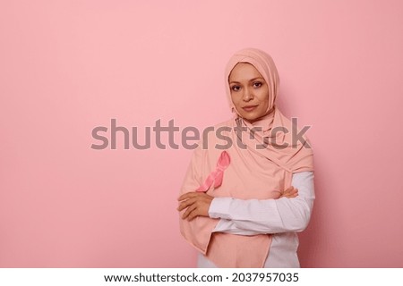 Confident portrait of gorgeous Middle Eastern ethnicity Arab woman, wearing pink hijab with pink Breast Cancer Awareness ribbon, looking at camera, posing over pink background with copy space