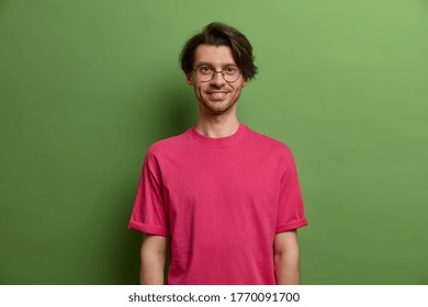 Confident Positive European Man With Happy Smile On Face, Has Dimples On Cheeks, Dressed In Bright Crimson T Shirt, Enjoys Day Off, Wears Big Optical Glasses, Models Against Green Background