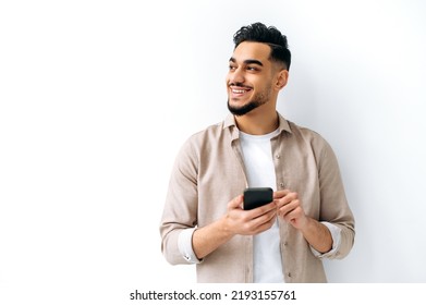 Confident positive handsome indian or arabian young man, holding smartphone in hand, chatting online, browsing internet, looking happily to the side, standing on isolated white background, smiling - Powered by Shutterstock