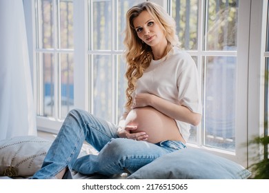 Confident pregnant swedish woman with wavy hair in white t-shirt and blue jeans sitting on the floor with pillows close to window, touching her belly, looking at camera.Woman in sunny day at home.