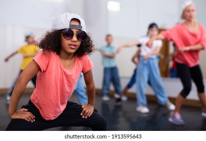 Confident preteen african american girl breakdancer in cap and sunglasses posing in dance studio with dancing children in background..